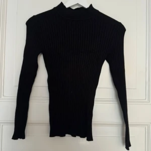 Svart ribbad polotröja - Säljer en svart ribbad polotröja med lång ärm och tight passform. Tröjan har hög hals och är tillverkad i ett stretchigt stickat material som sitter snyggt på kroppen. Perfekt att styla med jeans eller kjol för en clean look.
