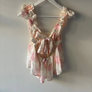Söt topp med öppen rygg från hm  - Supersöt blommig topp från H&M Divided med volangdetaljer och knyt i ryggen. Toppen är i ljusa toner av rosa och beige, har ärmlös design och ett luftigt, flowy material som känns perfekt för varma dagar. 