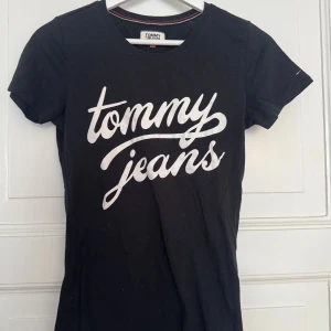 Svart t-shirt Tommy Jeans S - Svart t-shirt från Tommy Jeans i bomull med vit logga framtill. Klassisk rund halsringning och korta ärmar. Liten broderad flagga på ärmen. Perfekt för en avslappnad och stilren look.