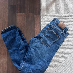 Blå jeans från Nudie Jeans - Säljer ett par klassiska blå jeans från Nudie Jeans med fem fickor och snygga kontrastsömmar. Jeansen har rak passform och är tillverkade i slitstarkt denimtyg av bomull. Modellen: ”Lean Dean”, storlek W32 L32