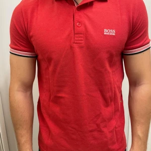 Röd pikétröja från Hugo Boss M - Snygg röd pikétröja från Hugo Boss med vit logga på bröstet. Tröjan har klassisk krage, knäppning framtill och ribbade ärmslut med svarta och vita detaljer. Regular fit och tillverkad i bomull för en skön känsla.