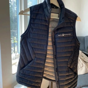 Mörkblå dunväst från Sail Racing - Snygg mörkblå dunväst från Sail Racing med horisontella sömmar och dragkedja framtill. Västen har logga på bröstet och är fodrad med Sail Racing Link Technology för extra komfort och värme. Perfekt att slänga över en hoodie eller tröja.  Priset kan diskuteras lägg ett bud! 