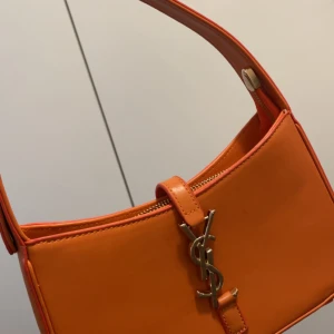 Orange axelväska från YSL i skinn - Säljer en snygg orange axelväska från YSL i slätt skinn med guldigt YSL-emblem framtill. Väskan har dragkedja upptill och röd insida. Perfekt storlek för det viktigaste och en riktig statement piece. Använt 1 gång 