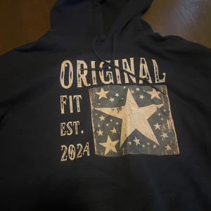 Mörkblå  hoodie med stjärntryck och text - Mörkblå hoodie med huva och snörning, tryck framtill med texten 'ORIGINAL FIT EST. 2024' och en stor stjärna omgiven av mindre stjärnor. Skön och avslappnad passform, perfekt för chill dagar. Storlek L