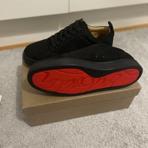 Christian Louboutin svarta sneakers - Snygga svarta sneakers från Christian Louboutin i mocka med klassisk röd sula och signaturdetaljer. Skorna har rund tå, svarta skosnören och en vit innersula med logotyp. Perfekta för dig som vill sticka ut med exklusiv streetstyle.