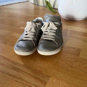 Gråa Saint Laurent sneakers i skinn - Snygga grå sneakers från Saint Laurent med klassisk låg profil och vit sula. Skorna är i mjukt skinn och har vita snören samt diskret logga på hälen och sulan. Perfekta för dig som gillar stilrena och exklusiva sneakers med en clean look. Endast en liten defekt på högersidan (syns knappt) därav priset
