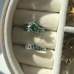 Silverringar med gröna stenar - Två matchande silverringar med glittrande gröna stenar. Den ena ringen har en större, oval grön sten i mitten och mindre stenar runtom, medan den andra har en vågig form med infattade små gröna stenar. 
