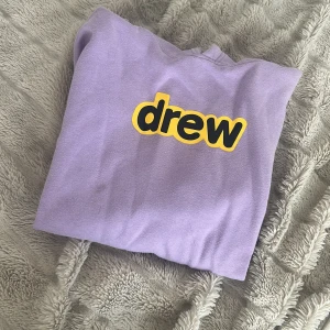 Lila hoodie från Drew House - Säljer en lila hoodie från Drew House med gul och svart logga på bröstet. Tröjan har huva, magficka och är i mjukt material som känns riktigt skönt. Perfekt för dig som gillar streetwear och vill sticka ut lite extra.