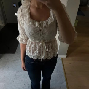 Spetsblus från Zara - Supersöt blus från Zara i ljusbeige spets med volangkantad urringning, knappar framtill och knytband i midjan. Blusen har trekvartsärmar med volang och är lätt transparent