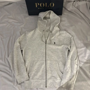 Grå hoodie från Polo Ralph Lauren - Snygg ljusgrå hoodie från Polo Ralph Lauren. Mjuk bomullsblandning och två fickor framtill. Perfekt för chill dagar eller när du vill ha en clean look. NYPRIS 2 395kr! Ralph lauren kassen ingår!