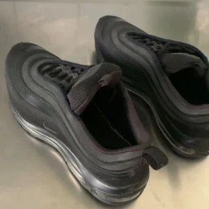Nike Air Max 97 Triple Black - Säljer ett par Nike Air Max 97 i helsvart färg med klassiska vågformade detaljer och reflekterande inslag. Skorna har snörning, platt sula och en ikonisk luftsula som ger grym komfort. Perfekta för dig som vill ha en clean och sportig look. Priset kan diskuteras!!