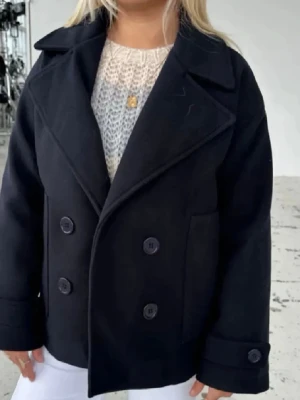 Svart kappa  - Stilren svart skepparkavaj med dubbelknäppning och breda slag. Jackan har stora knappar framtill och på ärmsluten, samt raka fickor på sidorna. Perfekt för dig som gillar klassisk och tidlös stil.