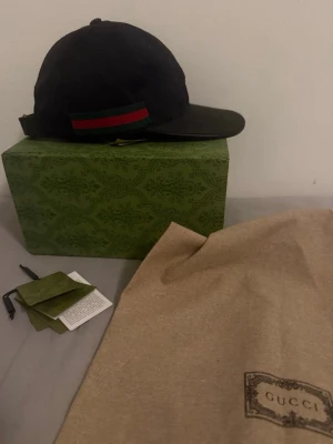 Svart keps med röd/grön detalj - Säljer en svart keps med klassisk röd och grön rand på sidan. Kepsen har justerbart spänne bak och är fodrad med svart tyg. 