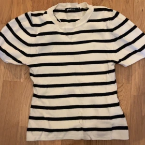 Randig topp från Gina Tricot - Kortärmad topp från Gina Tricot i vitt med svarta horisontella ränder. Modellen är figurnära med rund halsringning och ribbad kant. Perfekt till jeans eller kjol för en enkel och snygg look.
