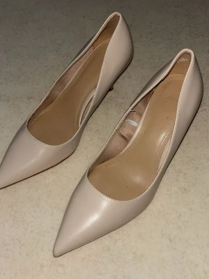 Beige pumps med spetsig tå från H&M - Säljer ett par stilrena pumps i beige från H&M. Skorna har spetsig tå och låg klack, vilket ger en elegant look. Tillverkade i syntetmaterial med slät finish. Perfekta för dig som vill ha en klassisk och enkel stil.