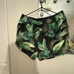 Svarta badshorts med grönt bladprint H&M - Snygga svarta badshorts från H&M med tropiskt bladprint i gröna nyanser. Shortsen har elastisk midja med snörning och innerbyxa i mesh. Perfekta för stranden eller poolen, riktigt fräsch vibe till sommaren.