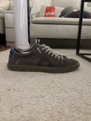 Grå oliver cabell  - Säljer ett par grå sneakers i mocka Oliver cabell med rund tå och platt sula. Skorna har ljusgrå snörning och en blå detalj på hälen. Snyggt cirkelmönster på sidan och diskret söm vid kanten. Perfekt för dig som gillar stilrena och klassiska sneakers.