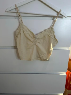 Beige spetsdetaljerad linnetopp - Säljer en ljusgul linnetopp med tunna axelband och fina spetsdetaljer längs urringningen. Toppen har en lätt rynkning framtill och är croppad i modellen. Väldigt mjukt och skönt material (tyget är en finare ljusgul i verkligheten)