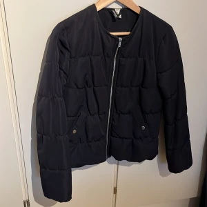 Svart pufferjacka från H&M Divided - Säljer en svart pufferjacka från H&M Divided i storlek 38. Jackan har quiltad design, rund halsringning och dragkedja framtill. Två fickor med tryckknappar och långärmad modell. Perfekt för kyliga dagar och enkel att matcha med olika outfits.