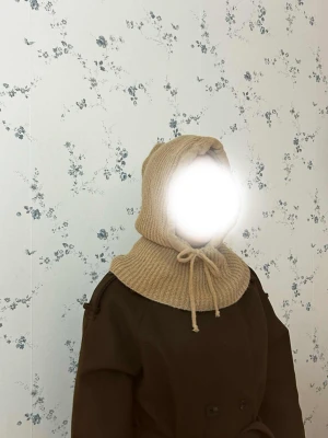 Beige stickad huvväst med snörning. Balaklava  - Säljer en beige stickad huvväst med ribbad struktur och snörning framtill. Västen har en bred krage som går upp över axlarna och en mysig huva. Perfekt för lager-på-lager och att hålla sig varm under kyliga dagar.