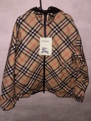 Burberry rutig vindjacka med huva - Säljer en snygg Burberry vindjacka med klassiskt rutigt mönster i beige, svart, vitt och rött. Jackan är vändbar med svart insida, har huva, dragkedja och dragsko vid ärmslut och nederkant. Perfekt för dig som vill ha en stilren och ikonisk look.