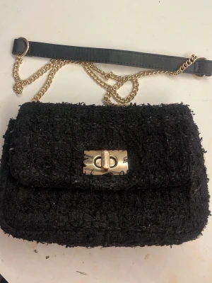 Svart axelväska med guld kedja - Säljer en liten svart axelväska i tweed-liknande material med guldfärgad kedjerem och snyggt vridlås i metall. 