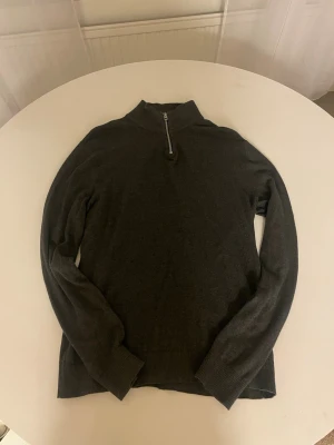 Mörkgrå quarter zip Jack & Jones - Snygg mörkgrå långärmad tröja från Jack & Jones med half zip i halsen. Tröjan har ribbade muddar vid ärmslut och nederkant, vilket ger en schysst passform. Perfekt för dig som gillar stilrena och enkla plagg med lite edge.