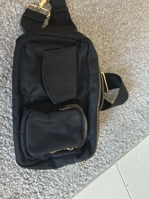Svart axelväska med gulddetaljer - Säljer en svart axelväska i syntetmaterial med bred axelrem och flera praktiska fack framtill. Väskan har snygga guldfärgade dragkedjor och detaljer, perfekt för att hålla koll på smågrejer. Enkel och stilren design som funkar till det mesta.