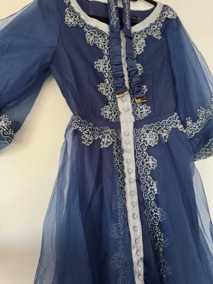 Marinblå långklänning med spetsdetaljer - Säljer en marinblå långklänning med transparenta ärmar och vackra vita spetsbroderier. Klänningen har markerad midja, dekorativa knappar framtill och puffiga ärmar. Materialet är lätt och ger en elegant känsla. Perfekt för dig som vill sticka ut med en unik look.