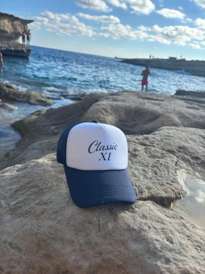 Classic X1 keps  - Vår marinblå keps med texten CLASSIC X1 kombinerar tidlös design med modern attityd. En perfekt accessoar för dig som vill ha en ren och självsäker look som passar både vardag och streetwear. Kepsen är tillverkad i slitstarkt material med bekväm passform och justerbar knäppning baktill för optimal komfort. Den marinblå färgen ger ett klassiskt uttryck, medan CLASSIC X1-trycket tillför en exklusiv och minimalistisk känsla  