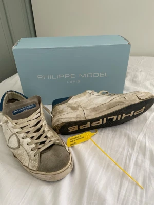 Philippe Model vita sneakers storlek 42 - Säljer ett par snygga Philippe Model sneakers i vitt skinn med grå mockadetaljer på tån och blå häl. Klassisk låg modell med snörning och svart sula där det står 'PHILIPPE MODEL' undertill. Perfekta till jeans eller shorts. 