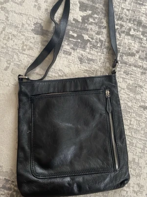 Svart axelväska i skinn - Säljer en stilren svart axelväska i skinn med justerbart axelband och en stor ficka framtill med dragkedja. Perfekt storlek för att få plats med allt du behöver och enkel att matcha till olika outfits.