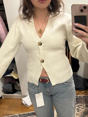 Vit kofta från Gina Tricot - Säljer en stilren vit kofta från Gina Tricot med två stora, mönstrade knappar framtill. Koftan har v-ringad hals, lång ärm och är gjord i mjukt stickat material. Passformen är normal och den är perfekt att slänga över en topp till jeans. Lite nopprig som de syns och pris kan diskuteras 
