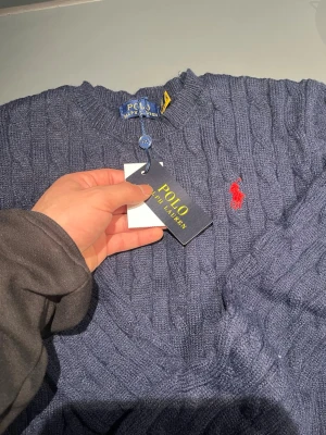 Blå stickad tröja från Polo Ralph Lauren - Säljer en marinblå stickad tröja från Polo Ralph Lauren med klassiskt kabelstickat mönster och röd broderad logga på bröstet. Tröjan har rund halsringning och ribbade muddar. Perfekt för dig som vill ha en tidlös och snygg look.
