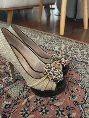 Beige pumps med leoparddetaljer - Unika pumps i beige textil med höga leopardmönstrade klackar och platåsula. Framtill finns en stor rosett i leopardmönster och repdetaljer. Skorna har öppen tå och svart kant runt öppningen. Perfekta för dig som vill sticka ut.