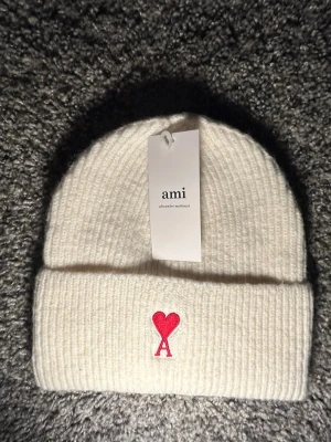 Vit stickad mössa från Ami - Vit ribbstickad mössa från Ami med röd broderad hjärtdetalj och bokstaven A framtill. Klassisk design med uppvikt kant, perfekt för kyliga dagar. Snygg och enkel att matcha med olika outfits.