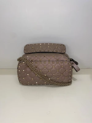 Valentino Garavani Rockstud Spike Free handväska - Snygg beige axelväska från Valentino med quiltat mönster och guldiga nitar. Väskan har kedjerem, dragkedja och ett stadigt handtag upptill. Tillverkad i mjukt skinn och har en lyxig vibe som verkligen sticker ut.