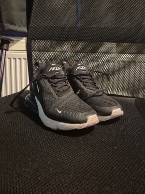 Nike Air Max 270 svarta sneakers - Svarta Nike Air Max 270 sneakers med vit sula och tydlig Air-enhet i hälen. Fick de i present förra året. Skorna har mesh-överdel för bra andningsförmåga, snörning och små vita Nike-loggor på sidan och tungan. Perfekta för dig som gillar sportig och clean stil. Är andvända bara några gånger. Kan tvättas innan de skickas!