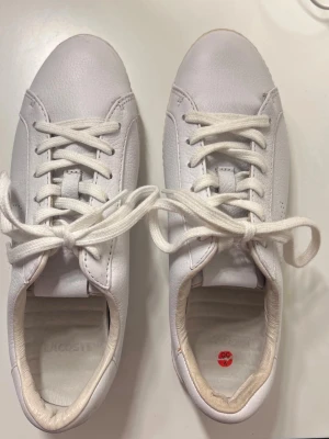 Vita sneakers från Lacoste - Snygga vita sneakers från Lacoste med klassisk design och rund tå. Skorna har snörning och är gjorda i skinn med en stilren, platt sula. Perfekta för dig som gillar en clean och fräsch look till vardags. Testade men aldrig blivit använda… dags att de får komma till användning 