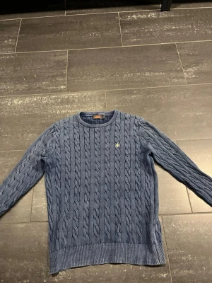 Blå kabelstickad tröja Morris - Snygg blå kabelstickad tröja från Morris med rund hals och diskret logga på bröstet. Tröjan har ribbade muddar vid ärmslut och nederkant, samt långärmad passform. Perfekt för dig som gillar klassisk stil med en modern twist.
