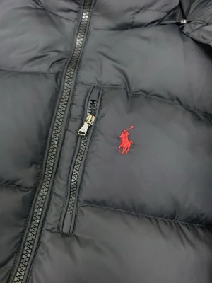 Svart pufferjacka från Polo Ralph Lauren - Säljer en svart pufferjacka från Polo Ralph Lauren med huva och dragkedja framtill. Jackan har en röd broderad logga på bröstet, flera fickor med dragkedja och justerbara detaljer vid ärmslut och nederkant. Perfekt för kalla dagar och riktigt snygg streetstil.