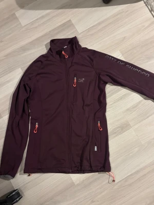 Vinröd vindjacka från 2117 of Sweden - vinröd/lila vindjacka från 2117 of Sweden med hög krage och tre fickor med dragkedjor och röda detaljer. Jackan har tryckt logga på bröstet och text på ärmen. (Ena är lite naggat, se bild) jackan är använd men har inga större eller avgörande tecken på slitage 