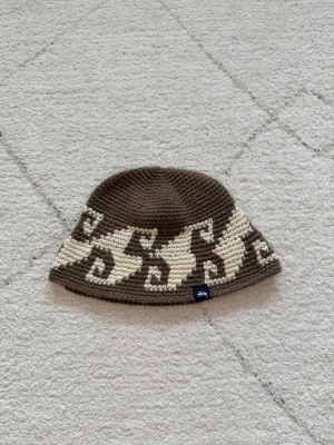 Brun och beige stickad Stüssy mössa - Säljer en brun och beige stickad mössa från Stüssy med ett unikt virkat mönster runt om. Mössan har en bucket hat-form och är tillverkad i tjockt garn som ger en cool retrovibe. Perfekt accessoar för dig som vill sticka ut lite extra.