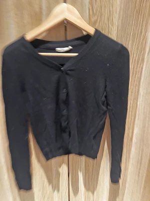 Svart kofta från KappAhl med knappar - En svart, klassisk kofta från KappAhl med rund halsringning och knäppning framtill. Långärmad modell i mjukt material som passar perfekt till lager på lager. Enkel och stilren design som funkar till det mesta.