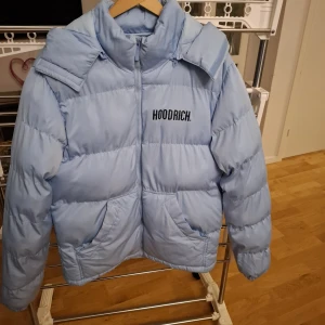 Ljusblå pufferjacka - Säljer en ljusblå pufferjacka från Hoodrich i storlek M. Jackan har hög krage, dragkedja framtill och två stora fickor. Hoodrich-loggan är tryckt på bröstet och insidan har coola detaljer med H-band. Perfekt för kalla dagar