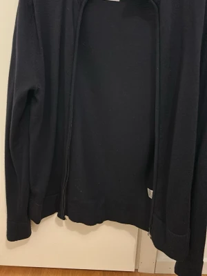 Svart kofta i merinoull med dragkedja - En enkel svart kofta i 100% merinoull med dragkedja framtill. Tröjan har långa ärmar och en rak passform. Perfekt att slänga på sig över en t-shirt. 