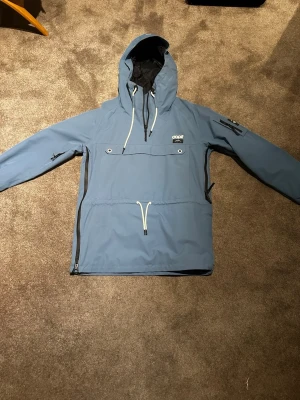 Blå vindjacka från Dope med huva - Säljer en blå skidjacka/annorak från Dope med stor huva och vita snören. Jackan har dragkedja i sidan, praktisk ficka på bröstet och ärmen samt justerbar dragsko i midjan. 