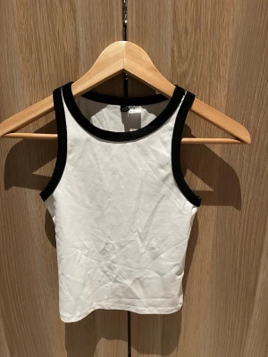 Vit och svart linnetopp Divided XS - Säljer en enkel och stilren linnetopp från Divided i storlek XS. Toppen är vit med svarta kanter runt hals och ärmhål, vilket ger en sportig kontrast. Tillverkad i mjuk bomull och har en tight passform som sitter snyggt på kroppen.