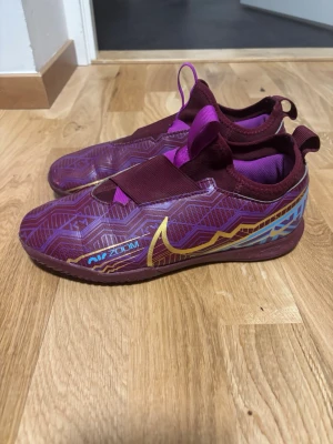 Nike Mercurial vinröda fotbollsskor - Säljer ett par Nike Mercurial fotbollsskor i vinrött med lila och blå geometriskt mönster och guldiga detaljer. Skorna har elastiskt band över vristen och dragflikar både fram och bak för enkel påtagning. Snygg design med Air Zoom-detalj på sidan.