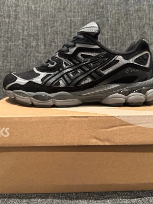 Svarta och grå Asics sneakers - Säljer ett par Asics sneakers i svart och grå med meshpaneler och detaljer i syntet. Skorna har en chunky sula och klassisk snörning. Perfekta för dig som gillar sportig stil och vill ha bekväma skor till vardagen.